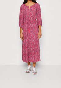 Femme portant une robe midi à manches longues et à fleurs roses, avec une ceinture à la taille, associée à des baskets blanches et roses, se tenant devant un fond uni.