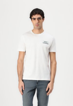 Camiseta de algodón blanca con cuello redondo y mangas cortas. Incluye un pequeño parche de logo en verde y negro en el lado izquierdo del pecho.