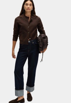 Cárdigan marrón con cuello alto, bolsillos frontales y botones plateados, combinado con jeans de mezclilla azul oscuro con puños enrollados y una chaqueta acolchada oscura.