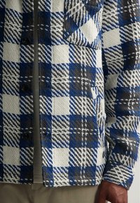 Gewoven blauwe, witte en zwarte gegadigde jas met een gestructureerde afwerking; twee knopen zichtbaar, vergezeld door een gelaagd grijs shirt eronder.