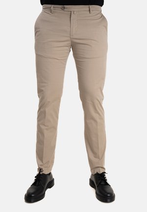 Chino beige con un taglio dritto, realizzati in un tessuto liscio. Presentano una chiusura con bottone e tasche frontali, indossati con scarpe nere.