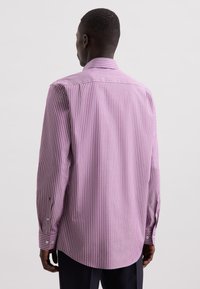 Mann trägt ein lila Langarmhemd mit weißen vertikalen Streifen, von hinten vor einem einfachen hellen Hintergrund gezeigt.