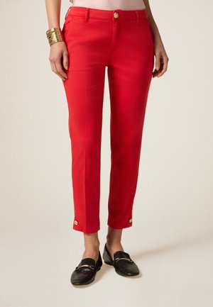 Pantalon en tissu coton rouge à coupe slim, avec une fermeture à bouton et des accents de bouton aux poignets. Porté avec des chaussures noires à enfiler.