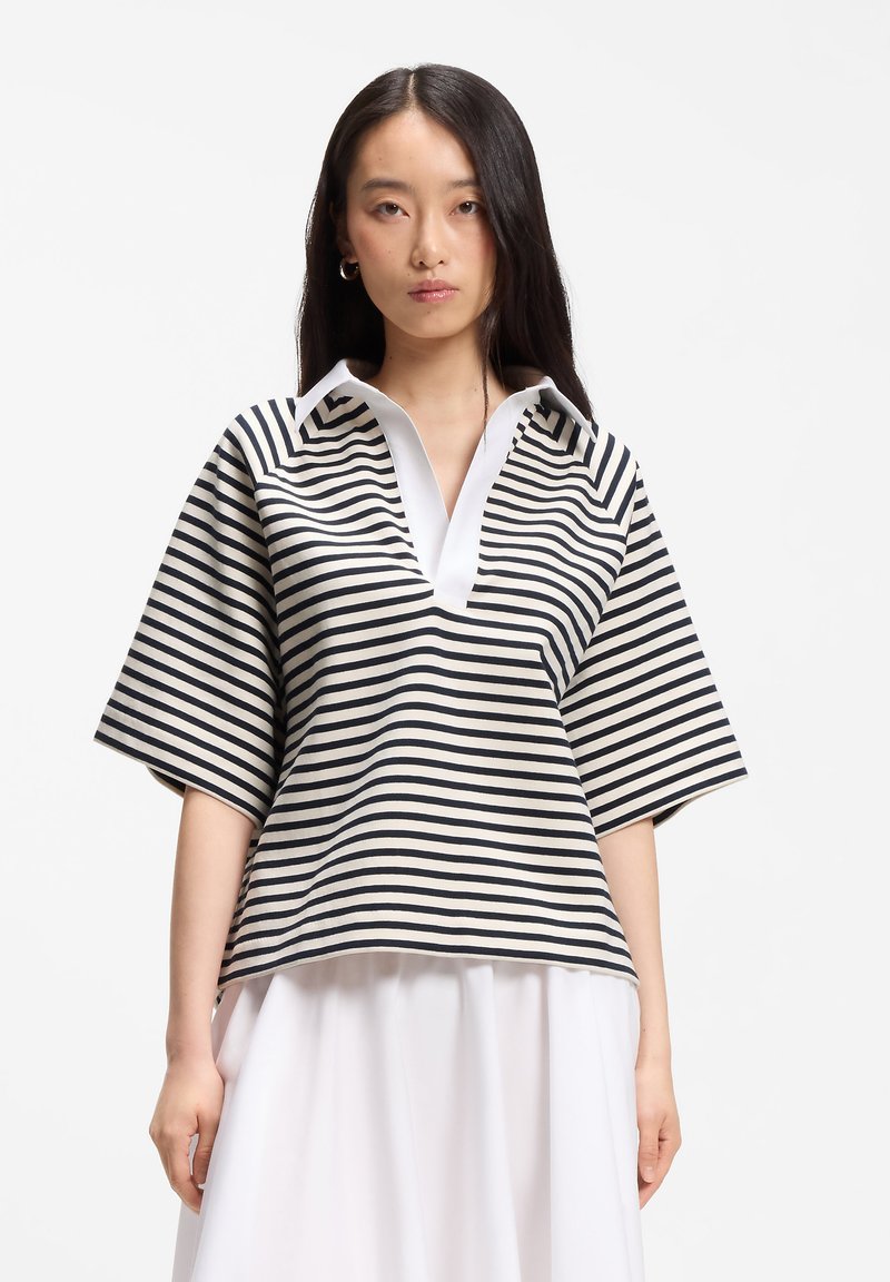 Femme portant un haut ample rayé horizontal noir et blanc avec un large col en V et une jupe blanche, debout devant un fond uni.