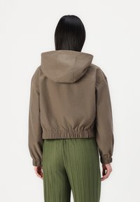 Chaqueta con capucha recortada en tela verde oliva con cinturilla y puños elásticos. Textura suave sin patrones visibles ni detalles de hardware.