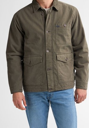 Man die een olijfgroene jas met knopen en drie voorzakken draagt over een lichtgrijs overhemd en een blauwe spijkerbroek, staand tegen een effen achtergrond.