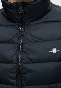 Gros plan d'une veste matelassée noire avec une fermeture éclair et un petit patch logo Gant sur la poitrine.