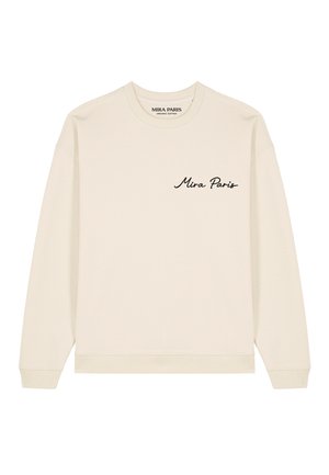 Blonde vrouw met krullend haar die een crèmekleurige trui draagt met "Mira Paris" in het zwart geborduurd op de borst.