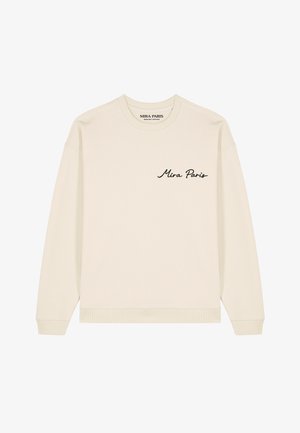 Femme blonde aux cheveux bouclés portant un sweat crème avec "Mira Paris" brodé en noir sur la poitrine.