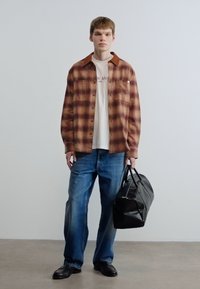 Καρό καφέ πουκάμισο, λευκό μπλουζάκι, μπλε τζιν, μαύρα παπούτσια, κρατώντας μια μαύρη τσάντα με σχέδια. Απλό φόντο, όρθια στάση.