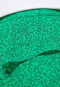 Grön blommig tyg med en texturerad yta, med en svart kontur av blommor i ett upprepande mönster. En något vikta kant syns.