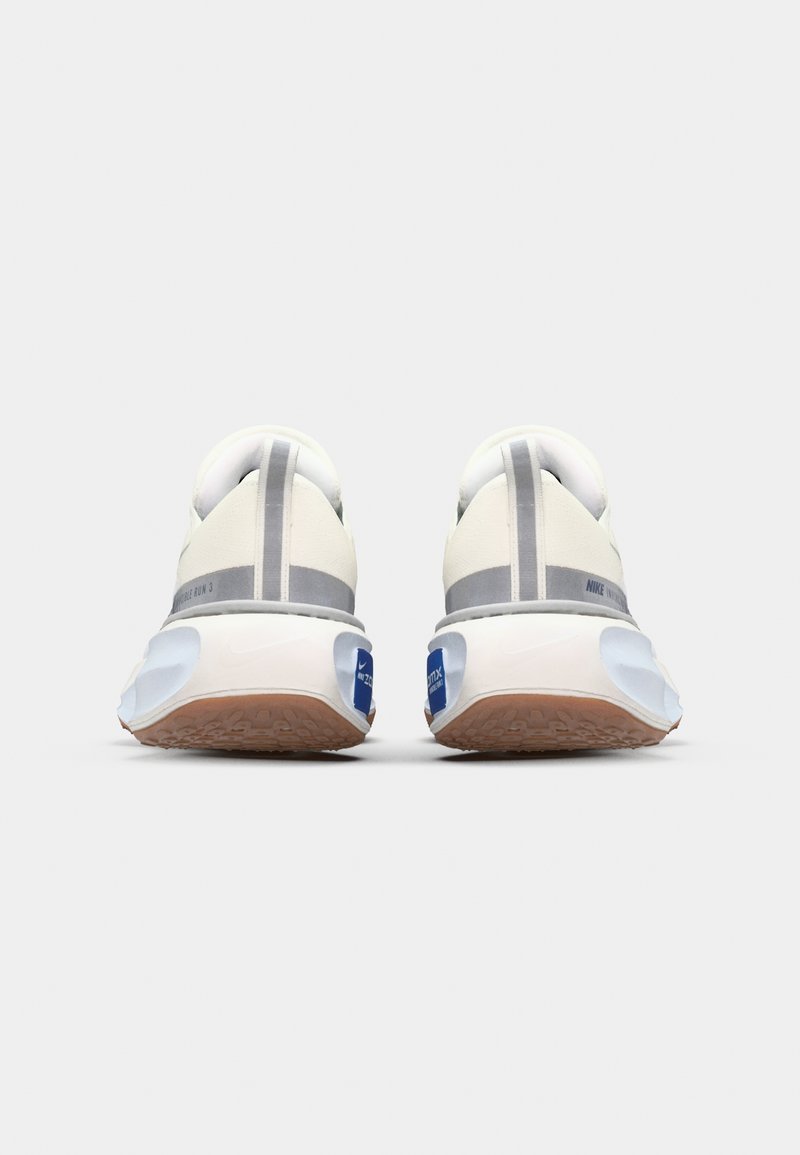 Zapatillas de correr Nike con una parte superior blanca suave, detalles en gris, una suela marrón texturizada y un logotipo azul en el talón. Diseñadas para la comodidad.