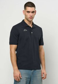 Mann står vendt mot kameraet iført en svart polotrøye med en hvit logo på brystet og lyseblå jeans mot en ensfarget bakgrunn.