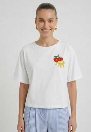 Femme aux cheveux foncés portant un t-shirt blanc avec des cerises brodées et le texte "oui", associée à un pantalon rayé bleu clair, souriant à la caméra.
