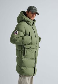 Grøn isoleret parka med hætte, der har lynlås og trykknaplukning, to forlommer og et logo-patch på venstre ærme.