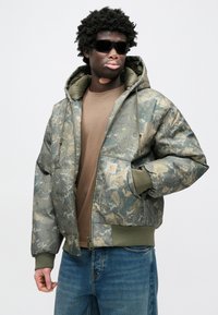 Maskáčová vatovaná bunda v zelených odstupoch s kapucňou, prednými vreckami a rebrovanými manžetami. Nosená cez hnedú košeľu a modré džínsy.