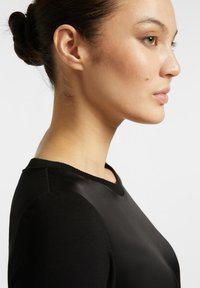 Černý top s kulatým výstřihem, který má hladkou texturu a přiléhavý střih. Má krátké rukávy a jemný lesk na látce.