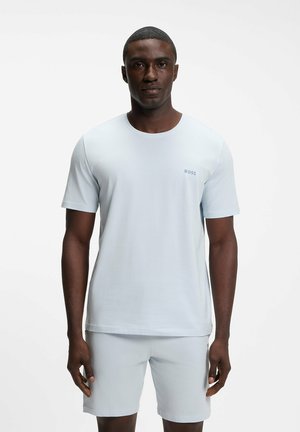 Mężczyzna stojący przodem, ubrany w jasnoniebieski t-shirt z krótkim rękawem i pasujące szorty na jednolitym białym tle.