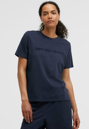 Frau mit lockigem Haar, die ein marineblaues T-Shirt mit umgekehrtem Text "SENSES THE LABEL" und passende marineblaue Hosen trägt.