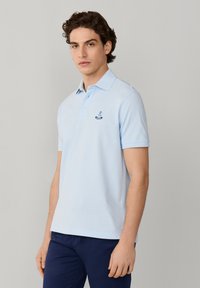 Młody mężczyzna z krótkimi kręconymi włosami, ubrany w jasnoniebieską koszulkę polo z małym logo oraz ciemnoniebieskie spodnie, stojący na tle jednolitego jasnego tła.