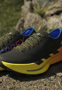Μαύρα και κίτρινα παπούτσια για τρέξιμο με μπλε εσωτερική επένδυση και κορδόνια με σχέδια που ακουμπούν σε μια βραχώδη εξωτερική επιφάνεια.
