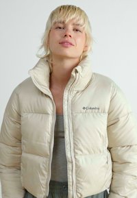 Doudoune beige avec col montant, fermeture éclair et motif piqué. Présente le logo "Columbia" sur la poitrine gauche.