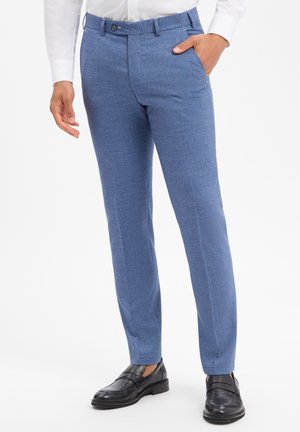 Mann trägt schlank geschnittene blaue Anzughose, schwarze Loafers und ein weißes Hemd mit Knöpfen, eine Hand in der Tasche, vor weißem Hintergrund.