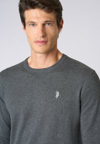 Maglione grigio lavorato a maglia con colletto rotondo e maniche lunghe. Presenta un logo ricamato discreto sul petto. Texture morbida e design aderente.