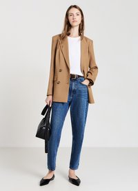Blazer marrone a doppio petto sopra una maglietta bianca, abbinato a jeans blu a vita alta e scarpe flat nere, mentre tiene una borsa nera.