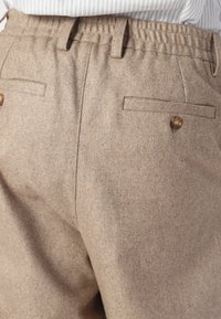 Beige Wollhosen mit einem elastischen Bund, zwei Gesäßtaschen und einem Knopfakzent. Der Stoff hat ein weiches, strukturiertes Aussehen.