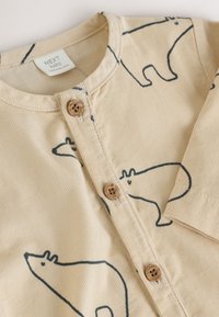 Beežidest sametriidest pusa, millel on tumesinised karu äärised, ees nööpkinnitusega puidust nööpidega ja voltidega varrukaserv.