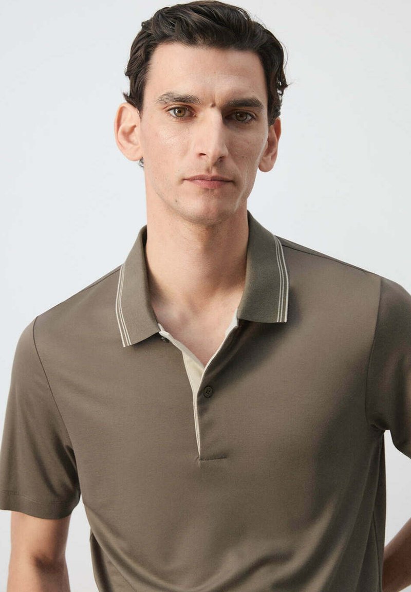 Man with short dark hair wearing a brown collared shirt with white trim stands against a plain light background, looking forward.