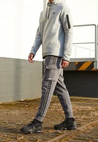 Grå Nike-hoodie med en zip-ficka och matchande grå joggers med en cargo-ficka och mönstrade sidostripes. Svarta sneakers fullbordar looken.
