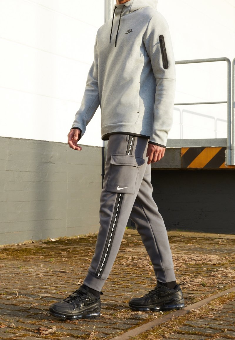 Grå Nike-hoodie med en zip-ficka och matchande grå joggers med en cargo-ficka och mönstrade sidostripes. Svarta sneakers fullbordar looken.