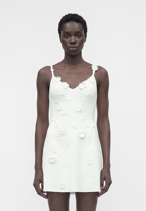 Fekete nő fehér ujjatlan miniruhában, 3D virágos hímzéssel, egyszínű világos háttér előtt, előre nézve áll.