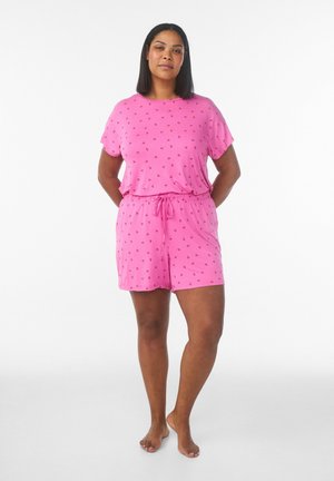 Donna in piedi a piedi nudi con le mani dietro la schiena, indossa un top rosa a maniche corte e shorts abbinati con una piccola stampa di ciliegie rosse.