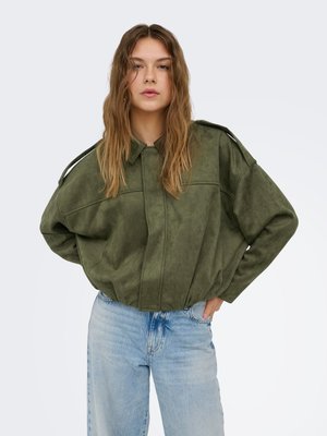 Jonge vrouw die een oversized olijfgroene jas met schouderbanden draagt en lichtblauwe hoge taille jeans, met handen in de zij.