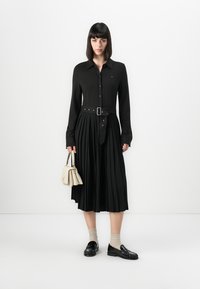 Svart langermet kjole med knapplukking foran og belte i midjen. Plisserte skjørtedesign. Modellen holder en kremfarget håndveske og bruker svarte loafers med sokker.