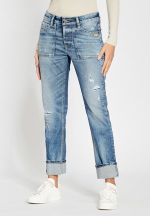 Kobieta w jasnoniebieskich jeansach z przetarciami i dużymi przednimi kieszeniami, z podwiniętymi nogawkami, beżowa bluzka z długim rękawem i białe sneakersy.