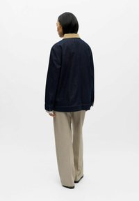 Giacca di jeans blu scuro con colletto beige, vestibilità comoda e maniche lunghe, abbinata a pantaloni larghi color chiaro.