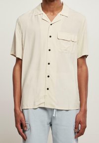 Homme portant une chemise beige à manches courtes avec une poche poitrine et un pantalon bleu clair avec des poches cargo.