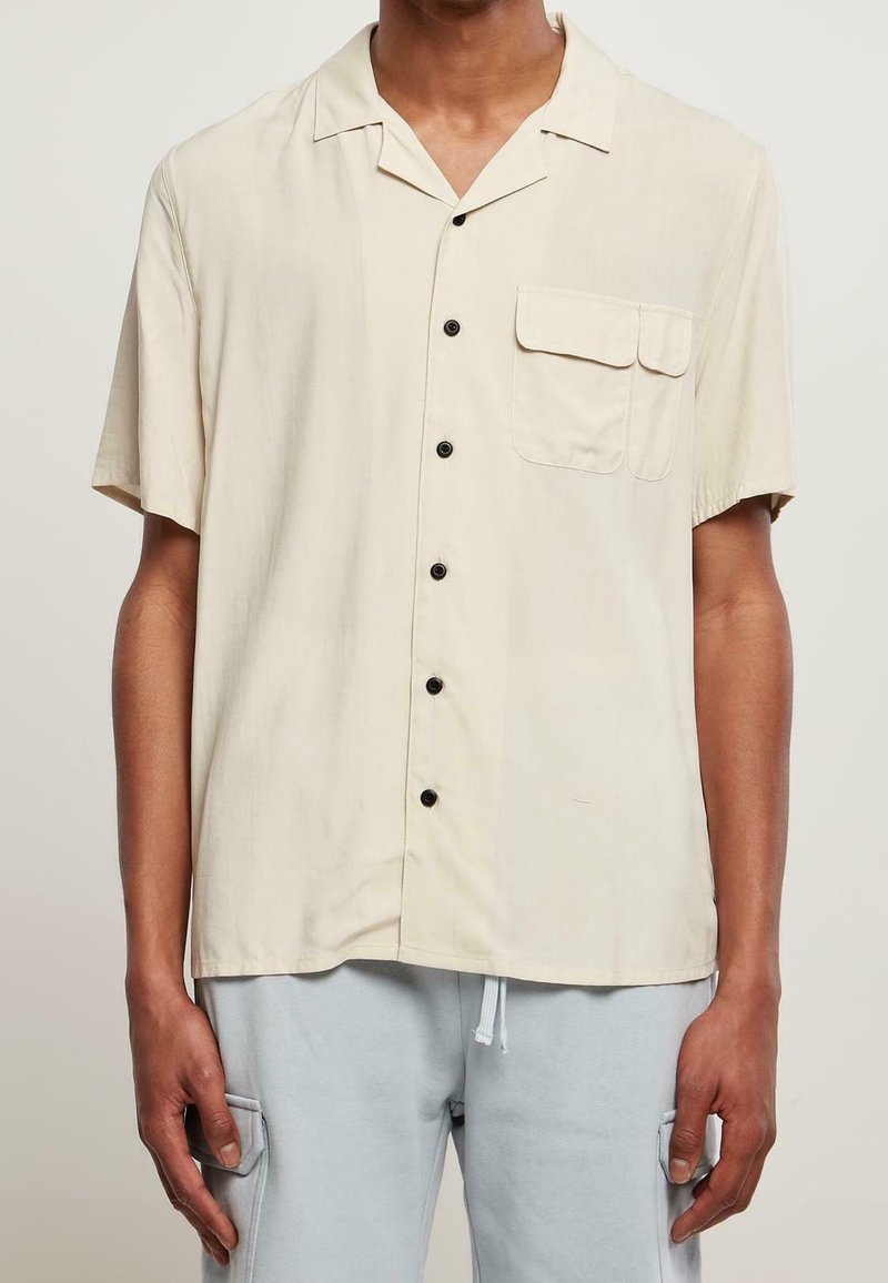 Homme portant une chemise beige à manches courtes avec une poche poitrine et un pantalon bleu clair avec des poches cargo.