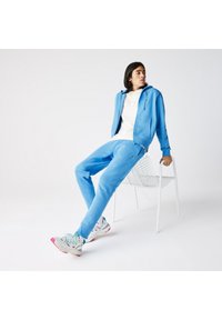 Blauwe katoenen rits-hoodie en bijpassende joggingsbroek gedragen met een witte polo. Grijze sportschoenen met roze accenten. Witte stoel op de achtergrond.