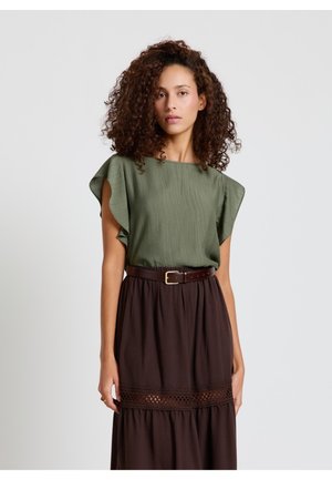 Frau mit lockigem Haar, die ein grünes Kurzarmoberteil trägt, das in einen dunkelbraunen Rock mit Spitzenverzierung und einem braunen Gürtel gesteckt ist, steht vor einem weißen Hintergrund.