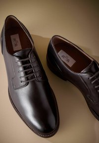 Zapatos de cuero marrón oscuro con un acabado suave, que presentan una puntera redondeada, cinco ojetes y costuras en contraste a lo largo de la suela.