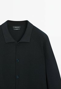Gebreide zwarte cardigan met een polokraag, twee knoopsluitingen en geribbelde textuur. Heeft raglanmouwen en een label met merknaam.