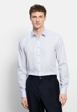 Jonge man draagt een lichtblauw gestreept overhemd met knopen, ingestopt in een zwarte broek, staande tegen een effen lichte achtergrond.