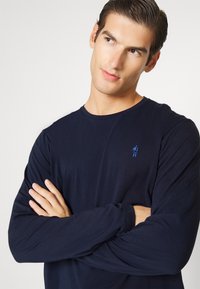 Marineblauw longsleeve shirt van gladde stof, met een klein blauw geborduurd logo op de borst. Eenvoudige ronde halslijn en aansluitende mouwen.