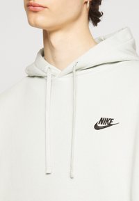 Harmaa huppari, jossa on nyörit ja musta brodeerattu Nike-logo. Pehmeä kangas ja minimalistinen muotoilu.