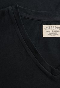 Czarny kołnierzyk koszulki z dekoltem w serek z beżową metką z napisem "SUPERDRY CO. Craft & Detail Trade Mark British Design" wszytą od środka.